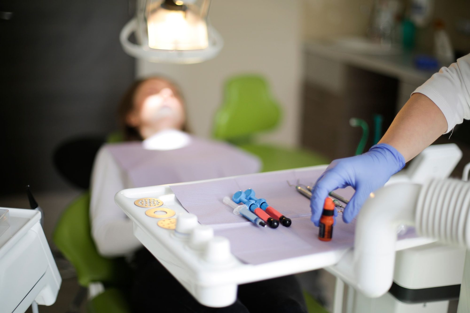 Medic cu mănuși care pregătește instrumentele pe tava de lângă scaunul stomatologic, cu pacienta în fundal